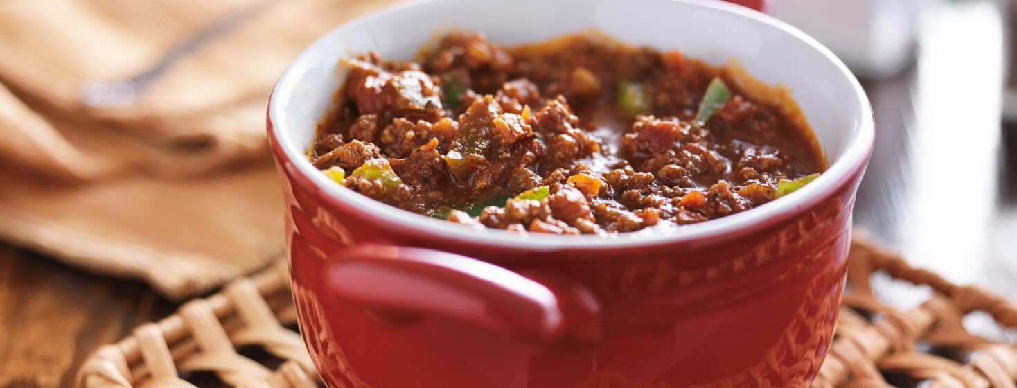 A bowl of chili with visible green pepper chunks sitting on a woven wicker pot holder in a red dutch oven.